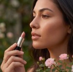 A woman with a glowing complexion applying a sheer soft bronze lip balm. The bottom section features the lip balm tube alongside four gold icons representing a radiant finish, lip hydration, organic ingredients, and a cruelty-free formula.
